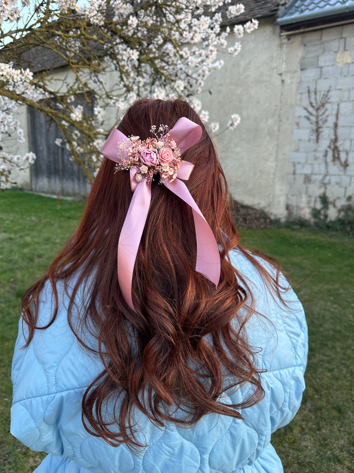 A long, wavy auburn-brown hair accessory featuring a light pink satin ribbon and a floral embellishment with pearls, shown worn on a person's hair in an outdoor setting.