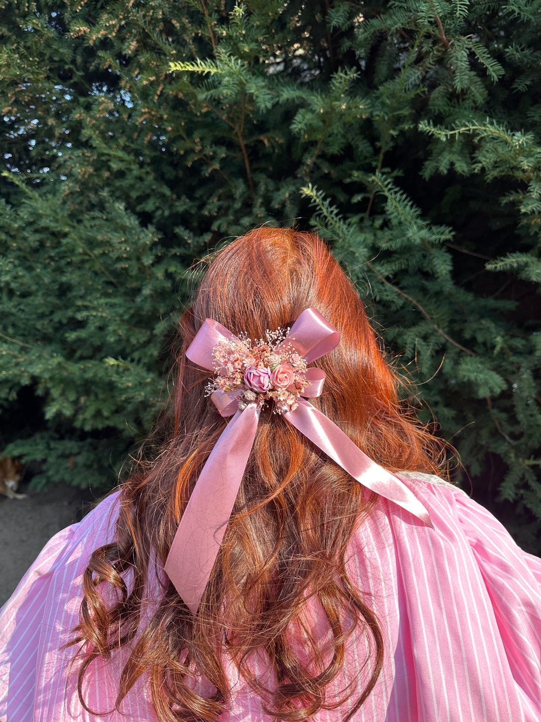 A pink satin ribbon hair bow with a floral center accessory, tied to a person's auburn hair, wearing a pink striped top against a green outdoor background.