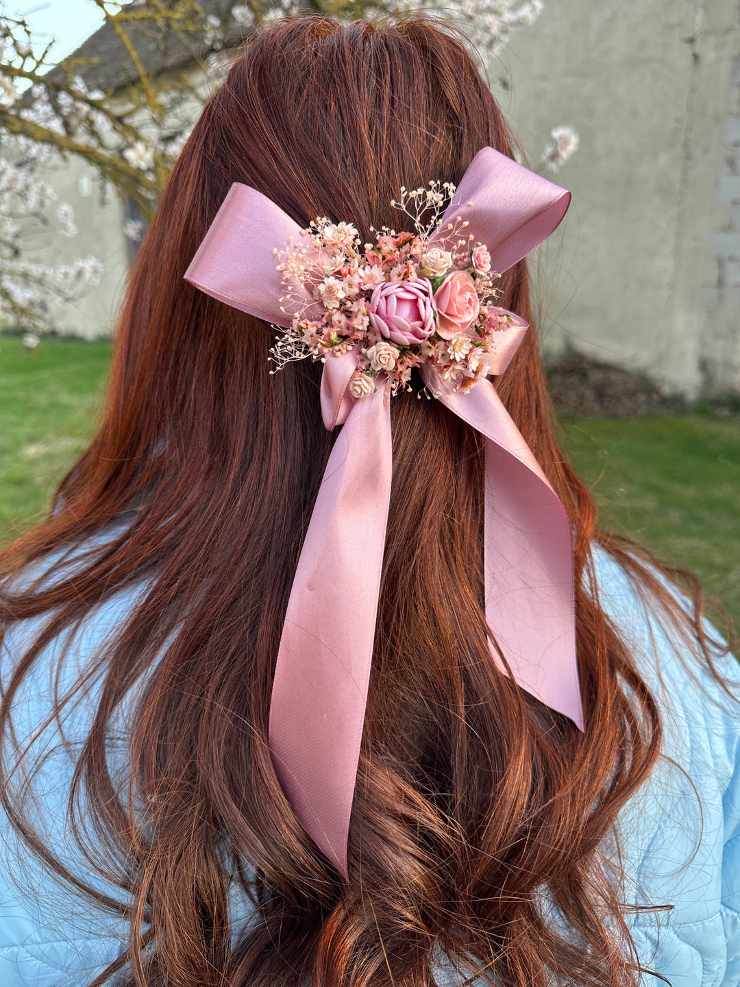 A long, wavy auburn-brown hair accessory featuring a large dusty pink satin bow and a central floral cluster with pink roses and dried flowers, shown worn on a person's head in an outdoor setting.