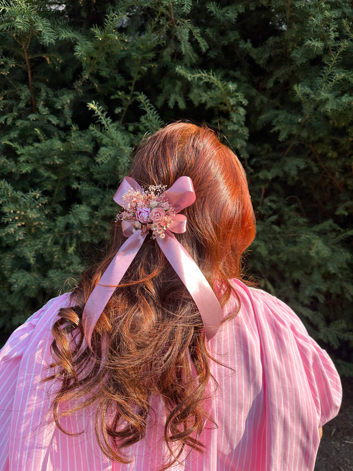 A close-up shot of a person's long, wavy auburn hair adorned with a light pink ribbon and a floral hair accessory, wearing a pink striped top against a background of green foliage.