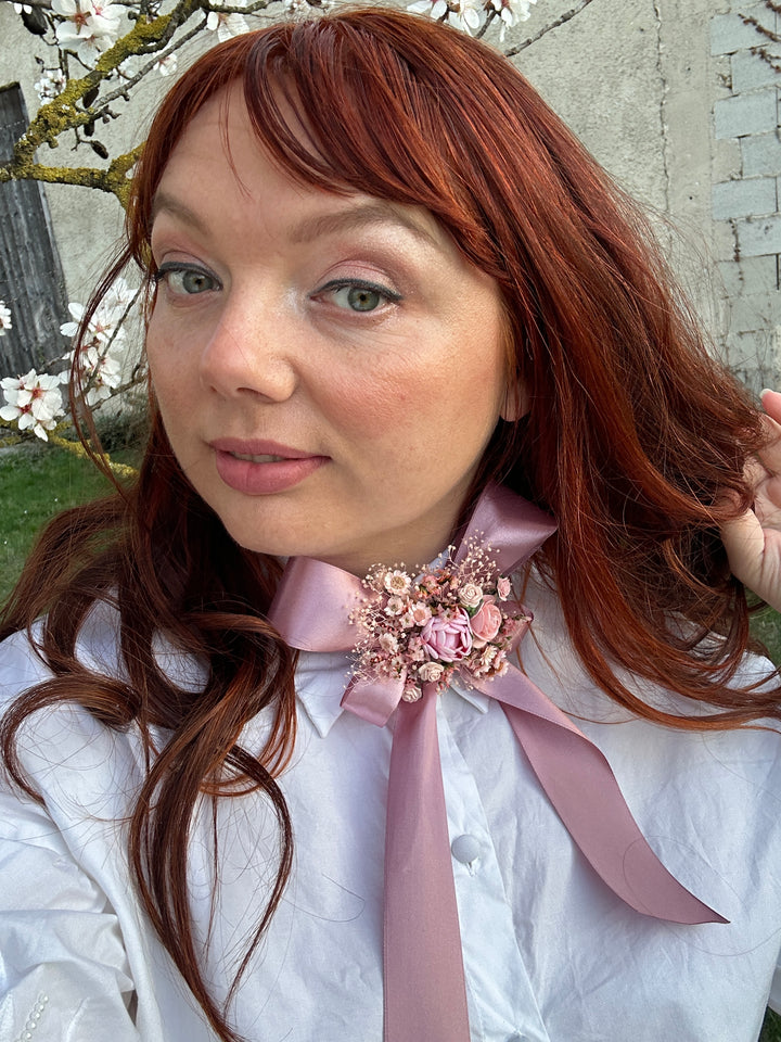 A close-up lifestyle shot of a woman with long red hair wearing a white shirt with a large dusty pink ribbon bow and a floral embellishment at the collar.