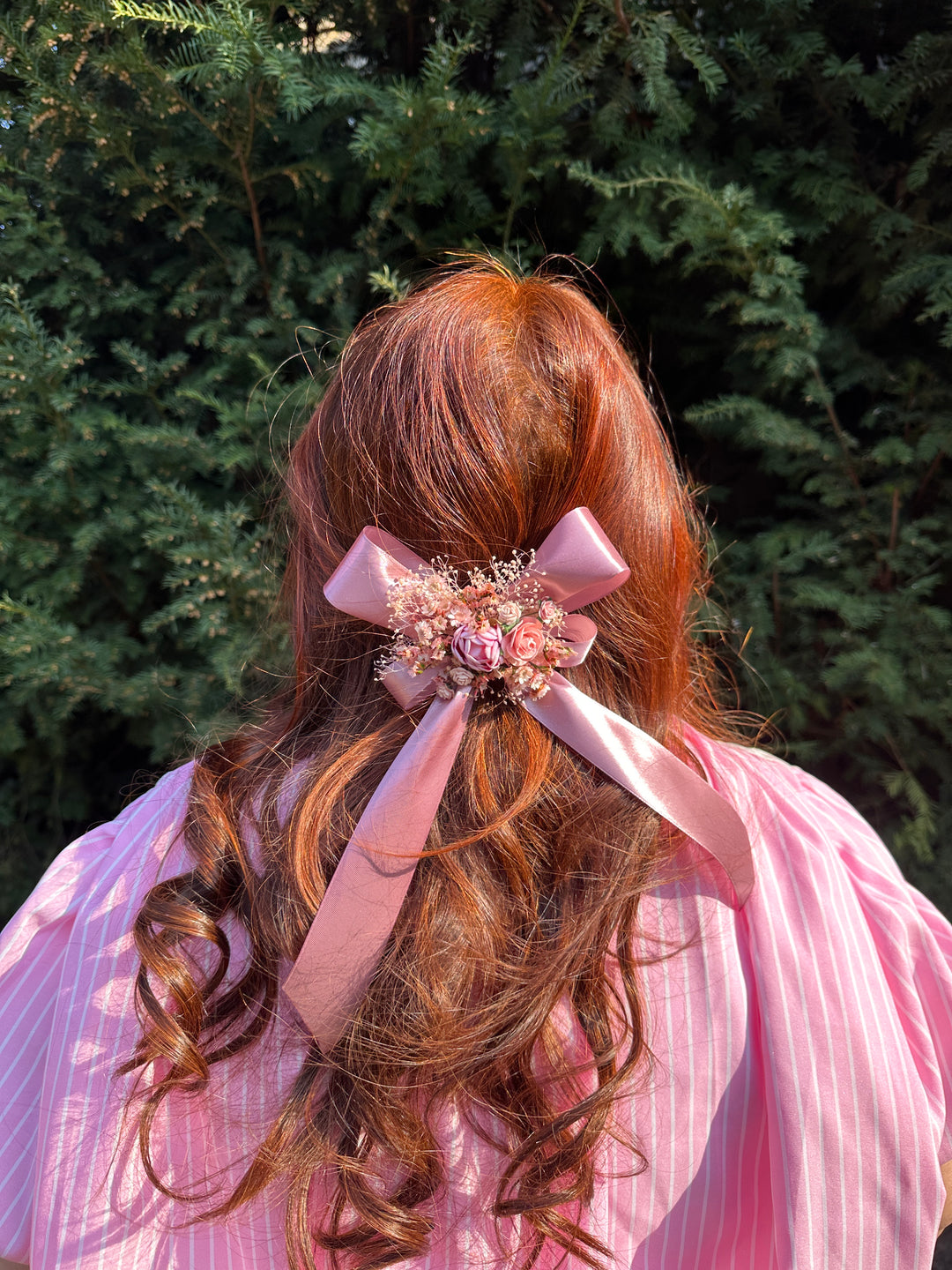 A pink satin ribbon hair bow with a floral center accessory, shown tied to a person's auburn hair outdoors against a dark green hedge.