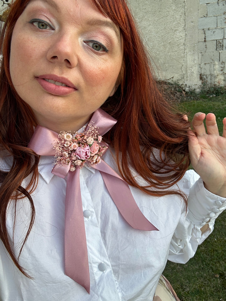 A close-up lifestyle shot of a woman with red hair wearing a white blouse with a large dusty pink ribbon bow and a decorative pink floral brooch at the collar.