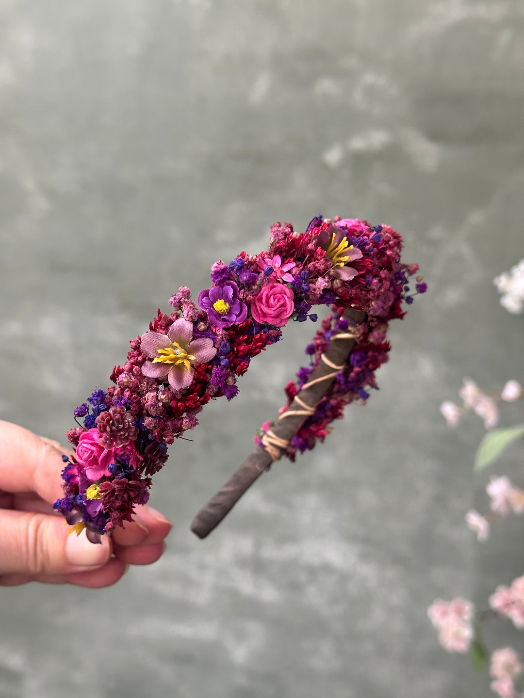 A hand holding a decorative headband covered in flowers with a gray textured background and blurred pink blossoms in the upper right.