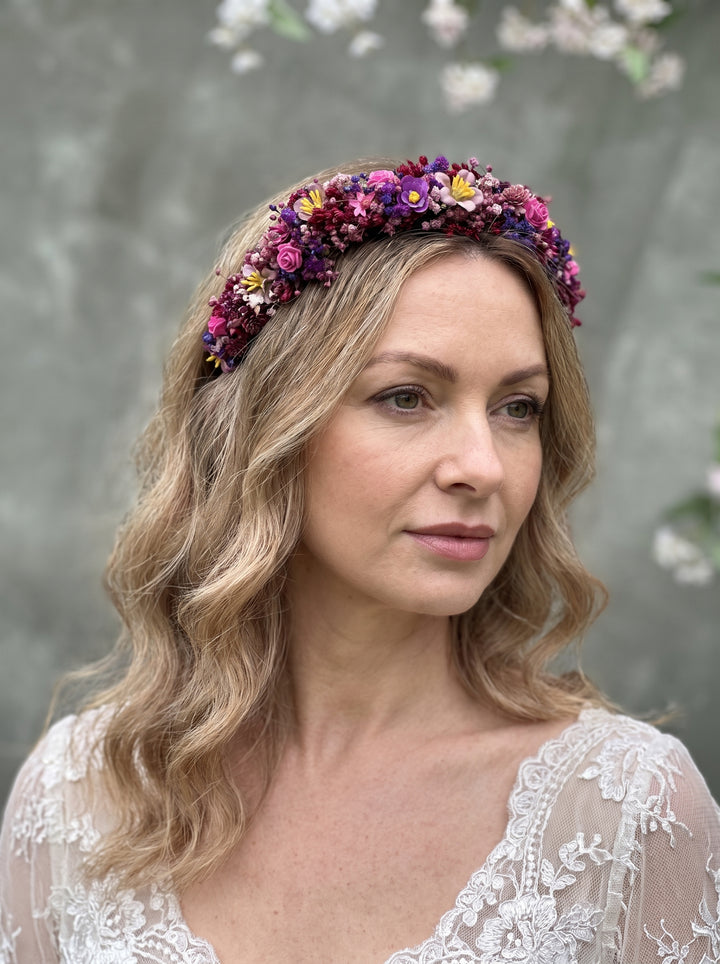 A woman with wavy blonde hair wearing a colorful flower crown headband and a white lace top, photographed against a soft gray background with blurred white blossoms.