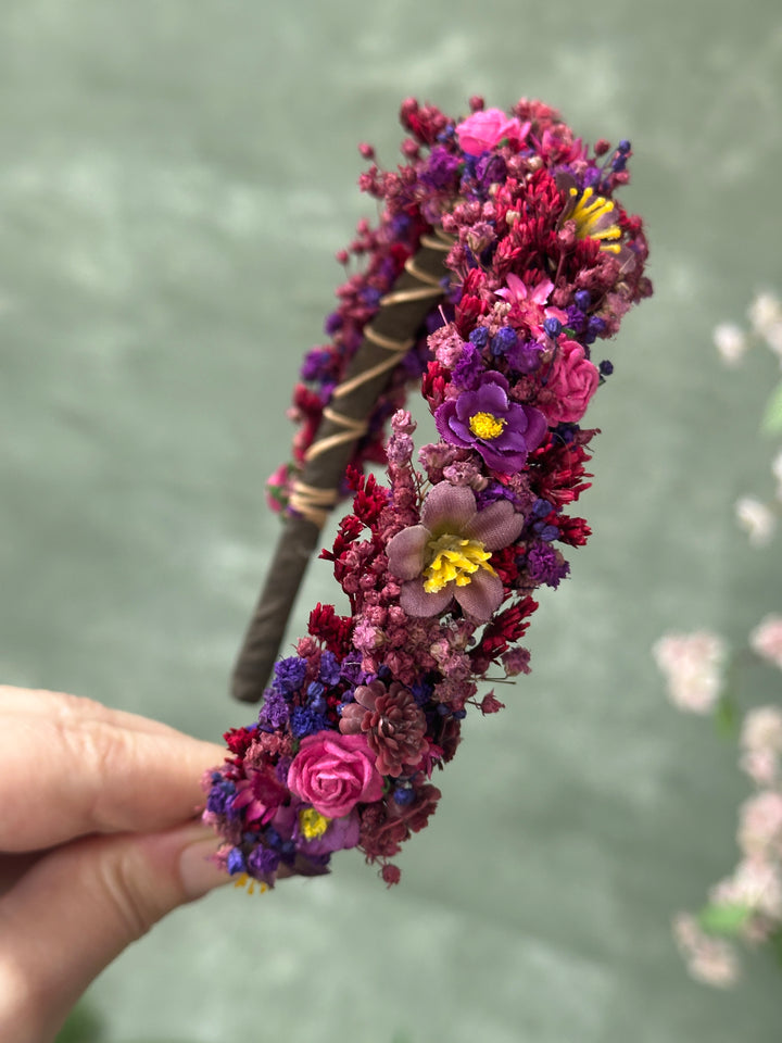 A hand holds a decorative headband covered in flowers in shades of pink, purple, and red, with small yellow centers and a brown band wrapped with thin gold wire, against a soft green blurred background.