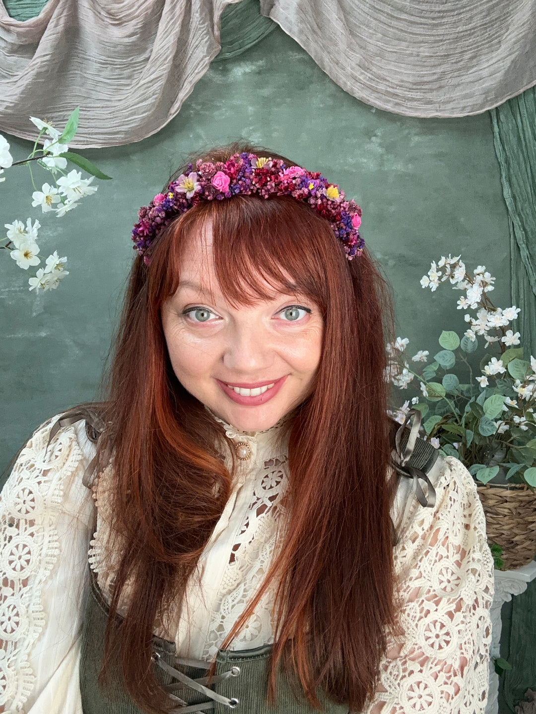 A smiling woman with long auburn hair wearing a colorful flower crown, dressed in a cream lace top, posed against a textured green background with floral props.