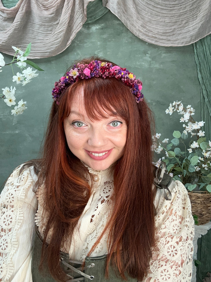 A smiling woman with long auburn hair wearing a colorful flower crown, dressed in a cream lace top, posed against a textured green background with floral props.
