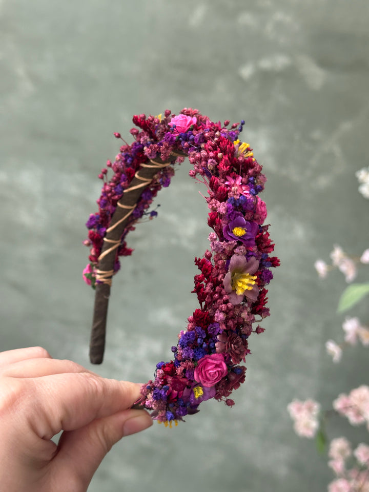 A hand holding a decorative headband covered in flowers and small wire accents, set against a soft gray background with blurred pink blossoms.