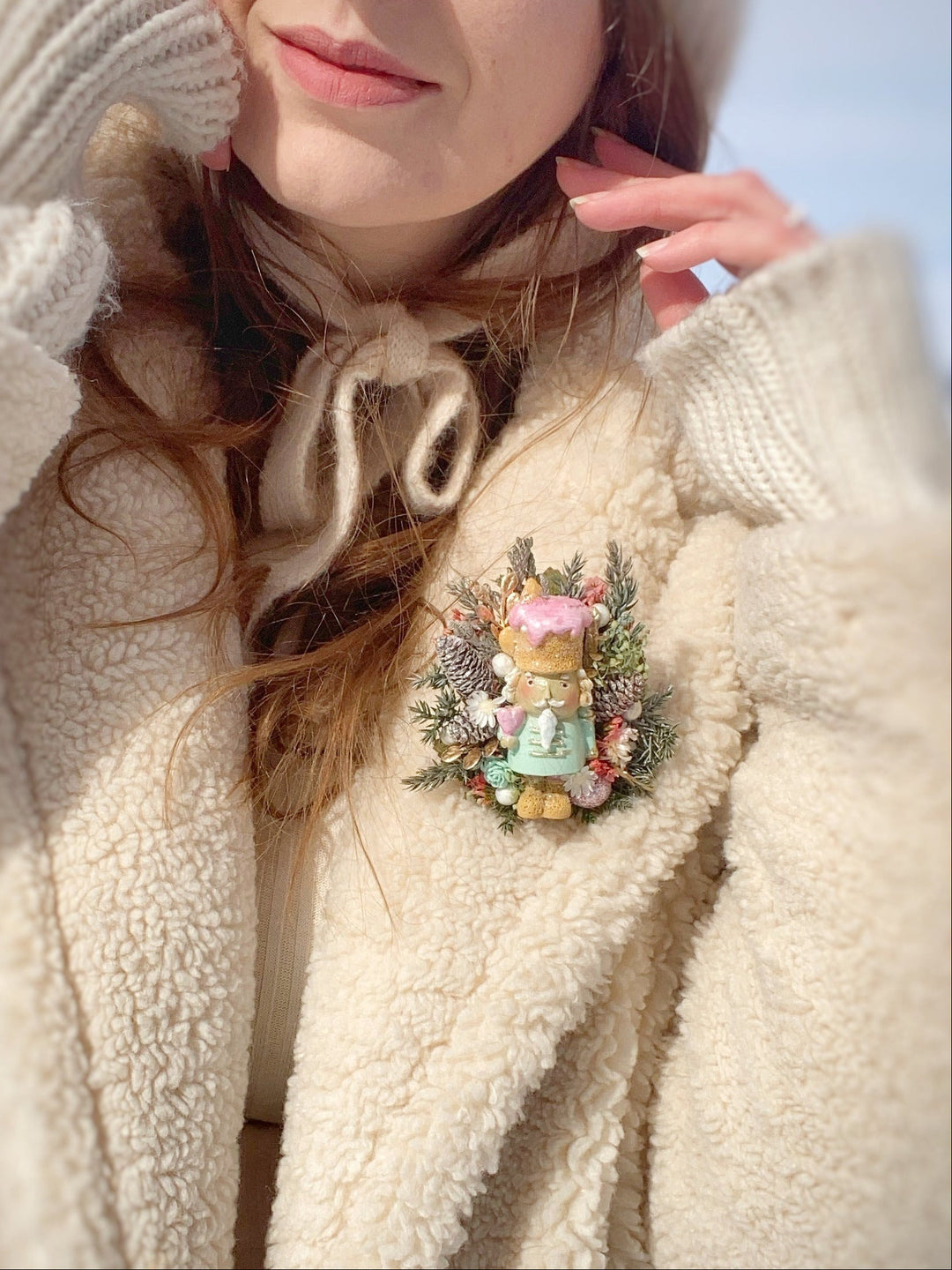 A close-up lifestyle shot of a woman wearing a cream-colored sherpa fleece coat, knit beanie, and matching knit gloves, featuring a decorative pastel teddy bear brooch on the chest.