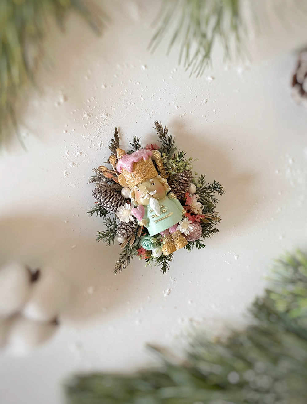 A small decorative brooch featuring a teddy bear figurine in a pink hat and green outfit, surrounded by pinecones, greenery, and white berries, photographed on a light cream surface with soft blurred holiday branches around the edges.