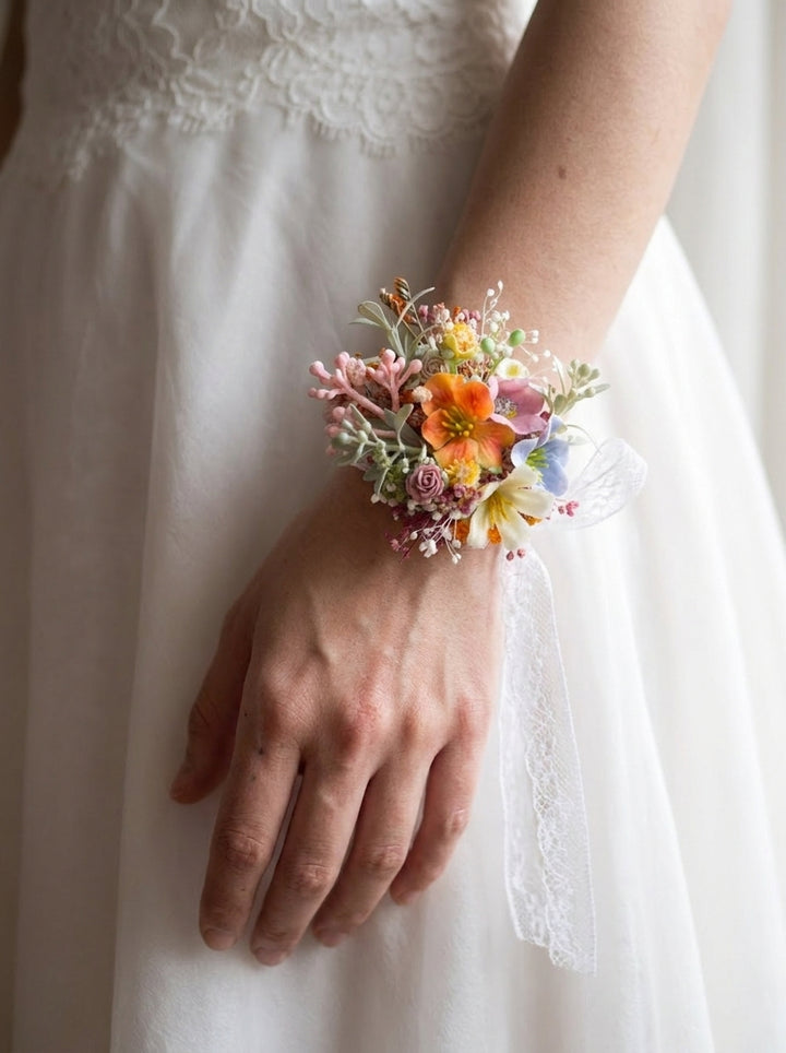 A close-up of a person wearing a white wedding dress, with a small wrist corsage featuring orange, pink, yellow, and blue flowers attached to the wrist.