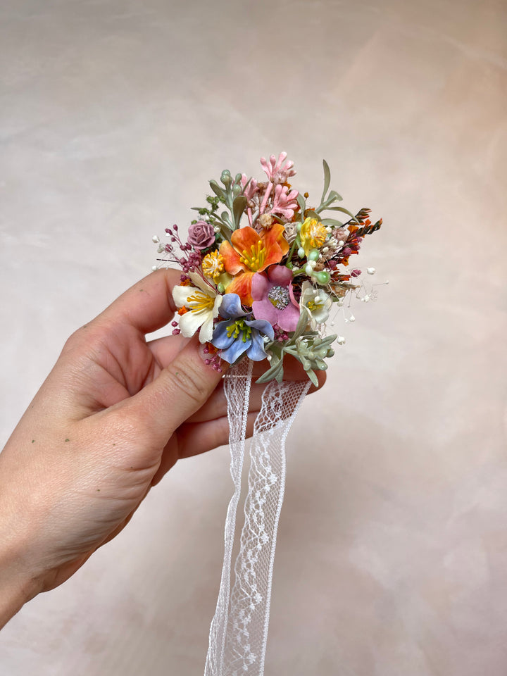 A hand holding a small floral corsage with a long white lace ribbon, set against a soft beige textured background.