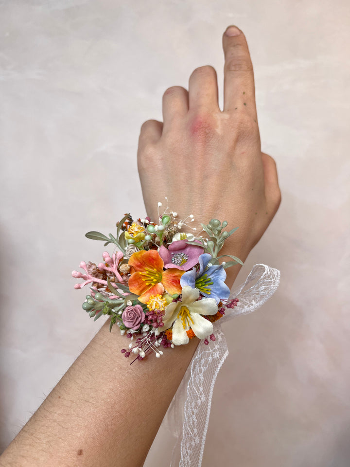 A wrist corsage with a delicate white lace ribbon, featuring a colorful mix of orange, blue, pink, and white flowers with green foliage, shown worn on a person's wrist against a soft beige background.