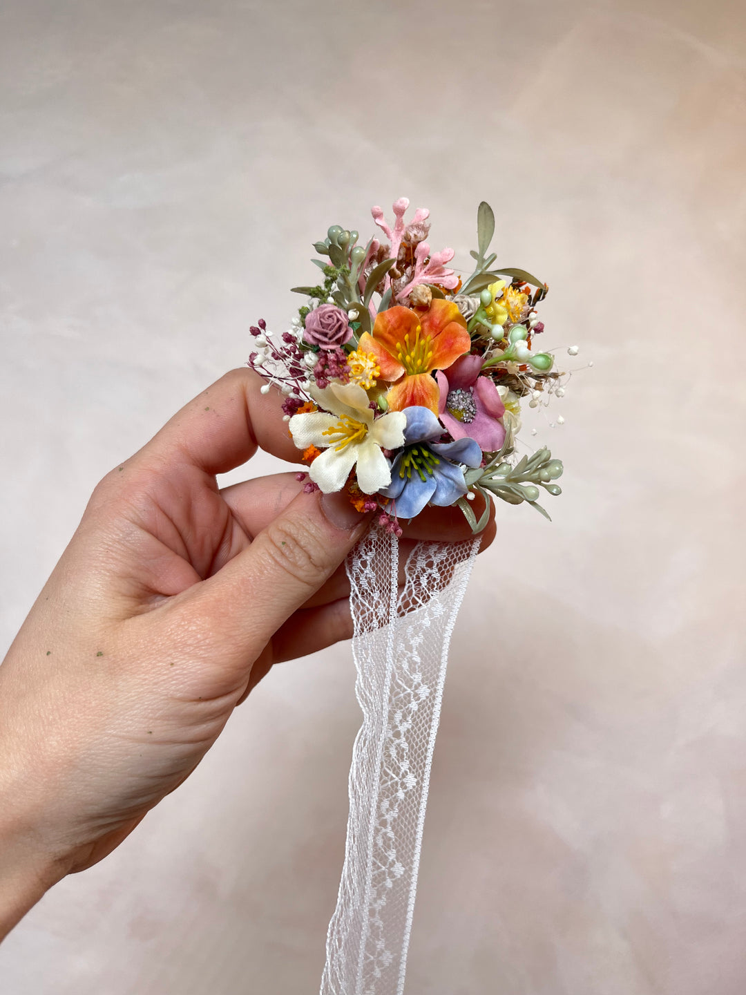 A hand holding a small artificial floral bracelet  with orange, pink, blue, and white flowers, green leaves, and a white lace ribbon, against a soft beige background.