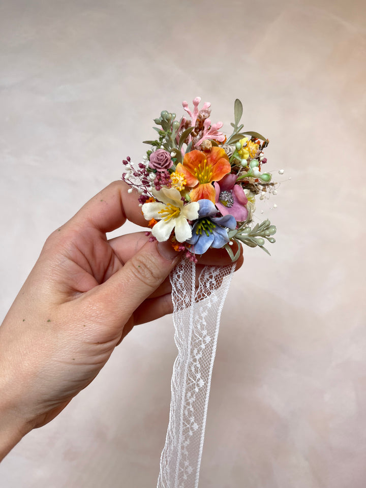 A hand holding a small artificial floral bracelet  with orange, pink, blue, and white flowers, green leaves, and a white lace ribbon, against a soft beige background.