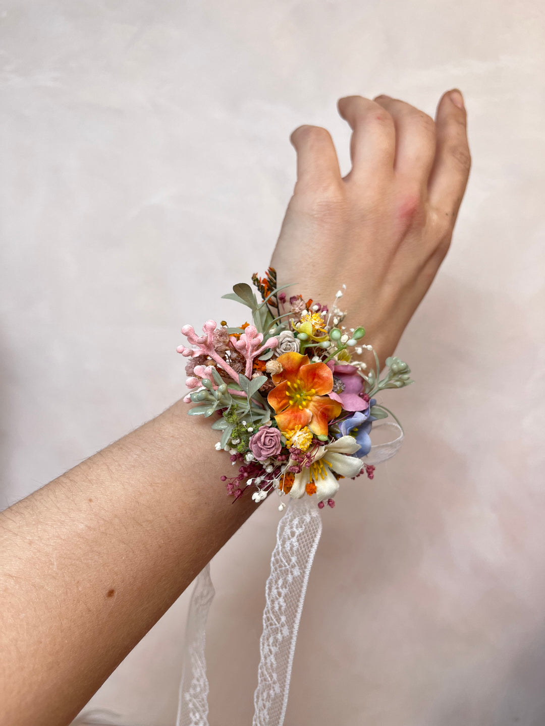 A close-up of a person's wrist wearing a small floral wrist corsage with orange, pink, purple, and yellow flowers, green leaves, and a white lace ribbon, against a soft beige background.