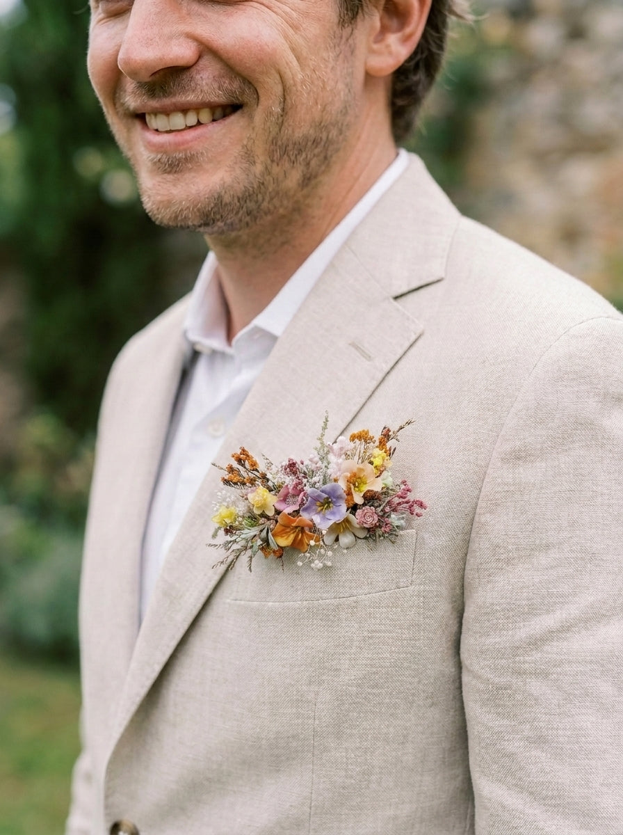 A man wearing a light beige linen suit jacket with a delicate multicolor wildflower boutonniere on his chest pocket, set against a soft, blurred outdoor garden background.