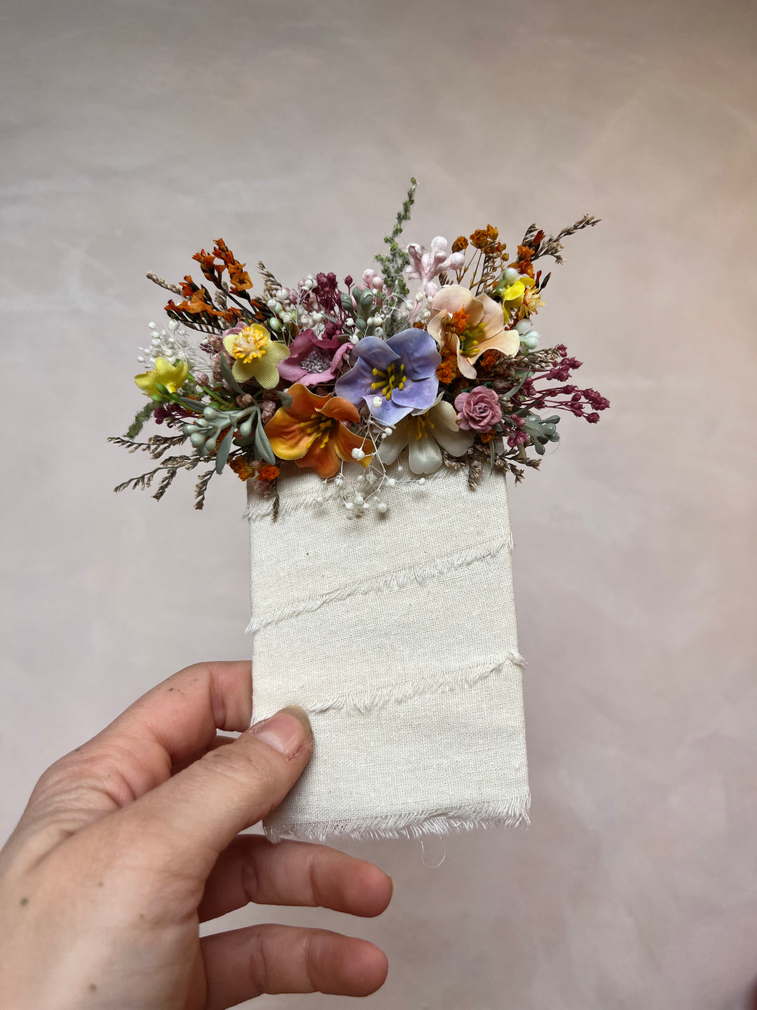 A hand holding a small  floral arrangement in orange, yellow, purple, pink, and green against a plain beige background.