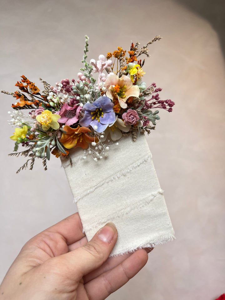 A hand holding a small dried flower arrangement attached to a square piece of off-white fabric, photographed against a soft beige background.