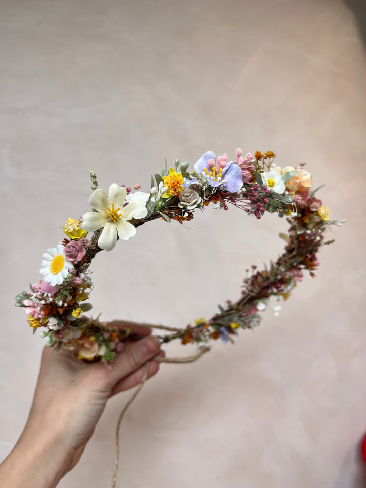 A hand holding a rustic floral crown made of flowers and greenery, including white daisies, small yellow blooms, lavender, and pink blossoms, against a soft beige background.