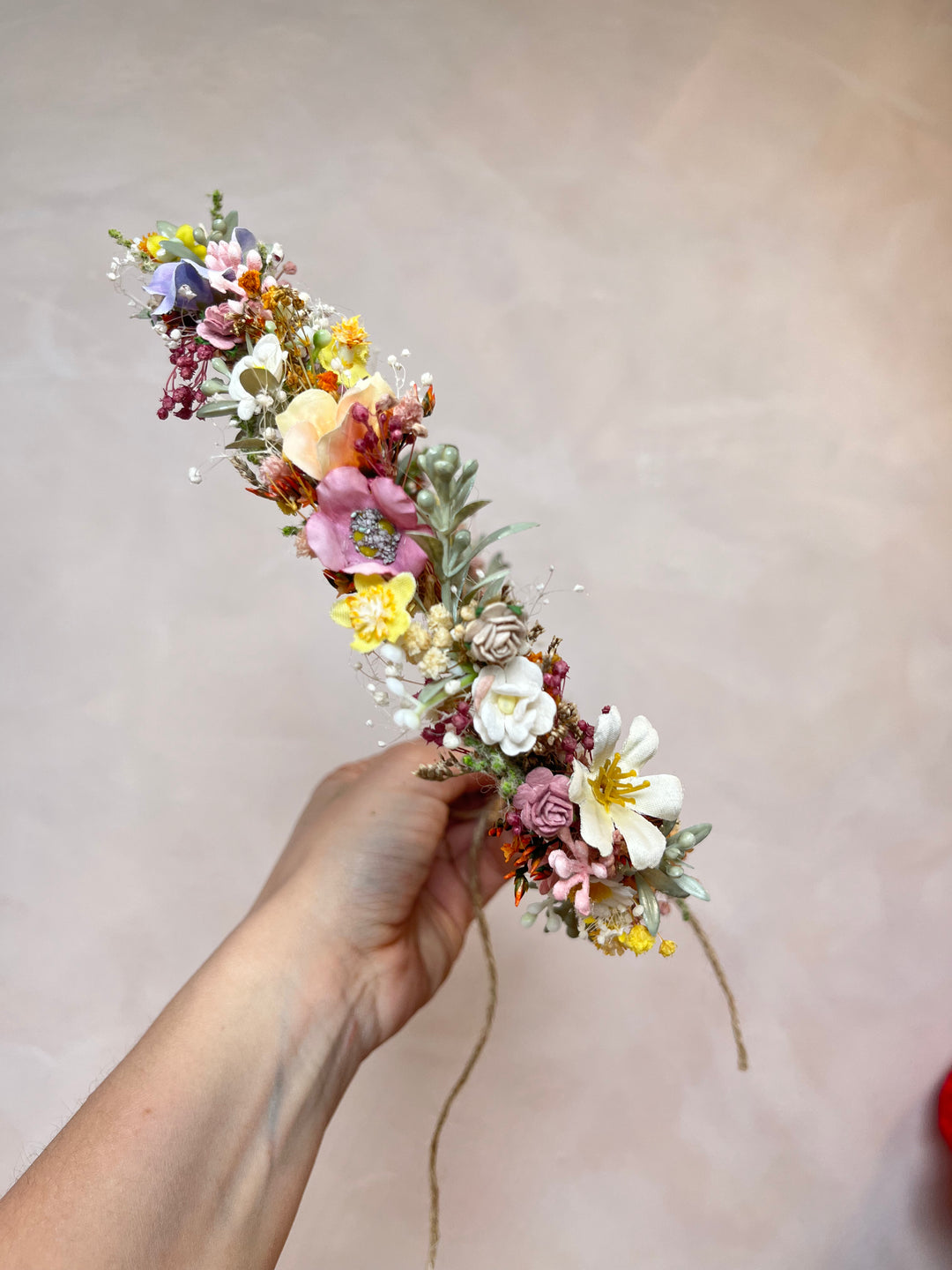 A hand holding a delicate flower crown made of pink, yellow, white, and purple flowers with green leaves, set against a soft beige background.