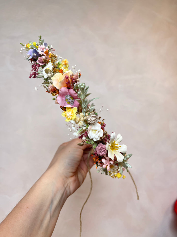 A hand holding a delicate flower crown made of pink, yellow, white, and purple flowers with green leaves, set against a soft beige background.