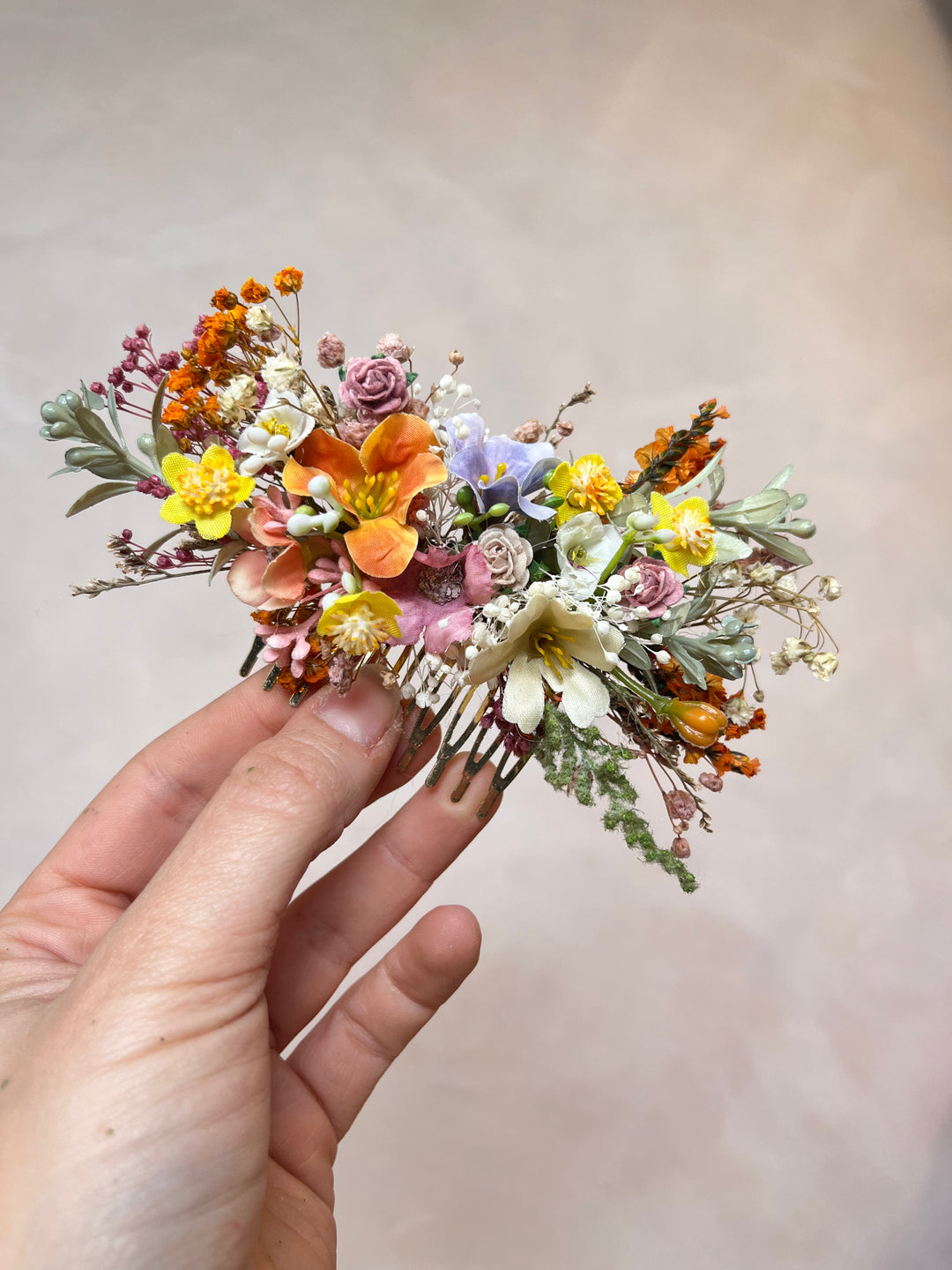 A hand holding a small dried flower hair comb accessory with orange, yellow, pink, white, and green botanical elements against a soft beige background.