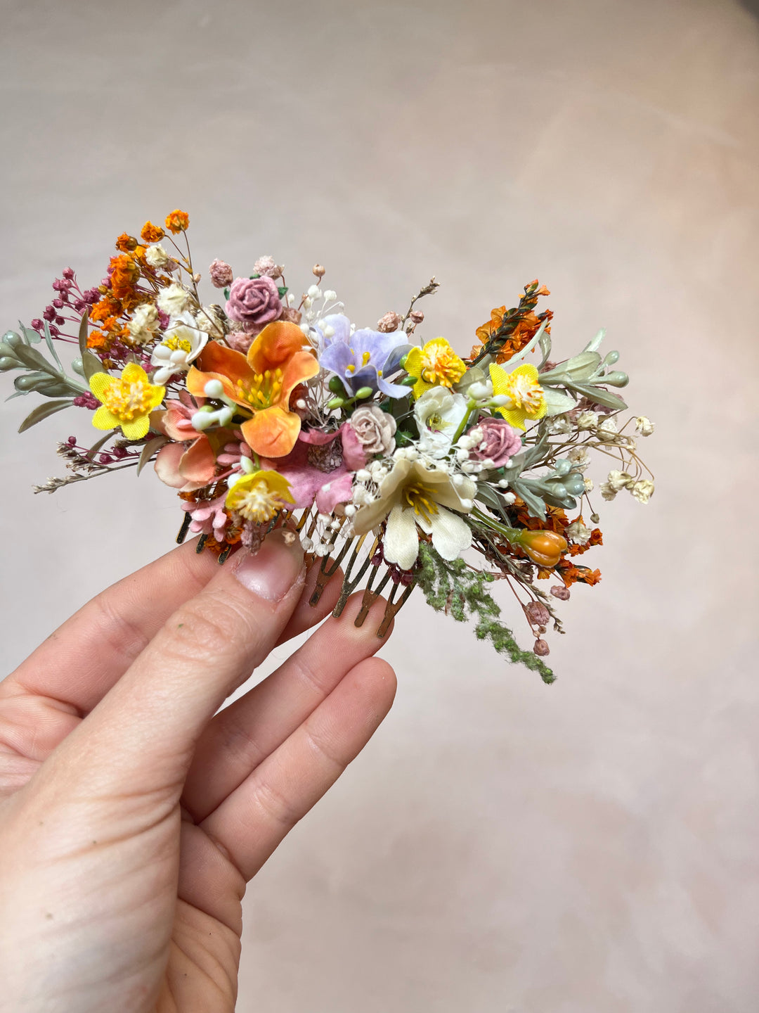 A hand holding a small floral hair comb featuring dried orange, yellow, pink, and white flowers with green leaves, set against a soft beige background.