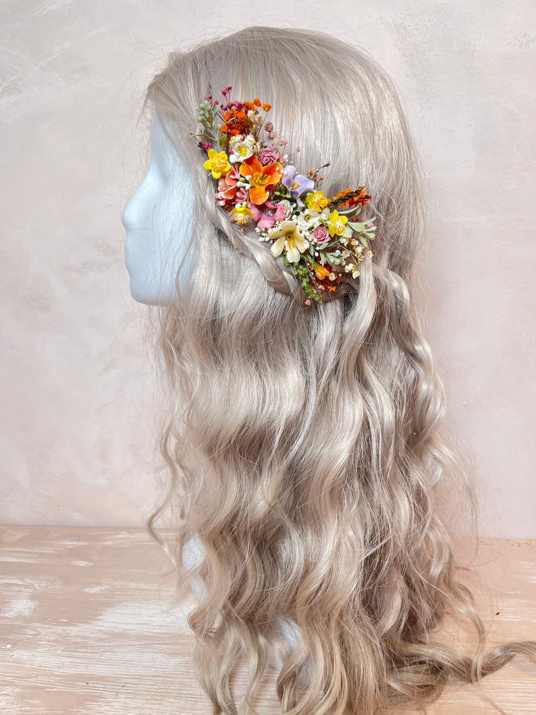 A long wavy platinum blonde wig displayed on a mannequin head in profile, decorated with a colorful floral hair accessory near the crown, against a soft beige textured background.