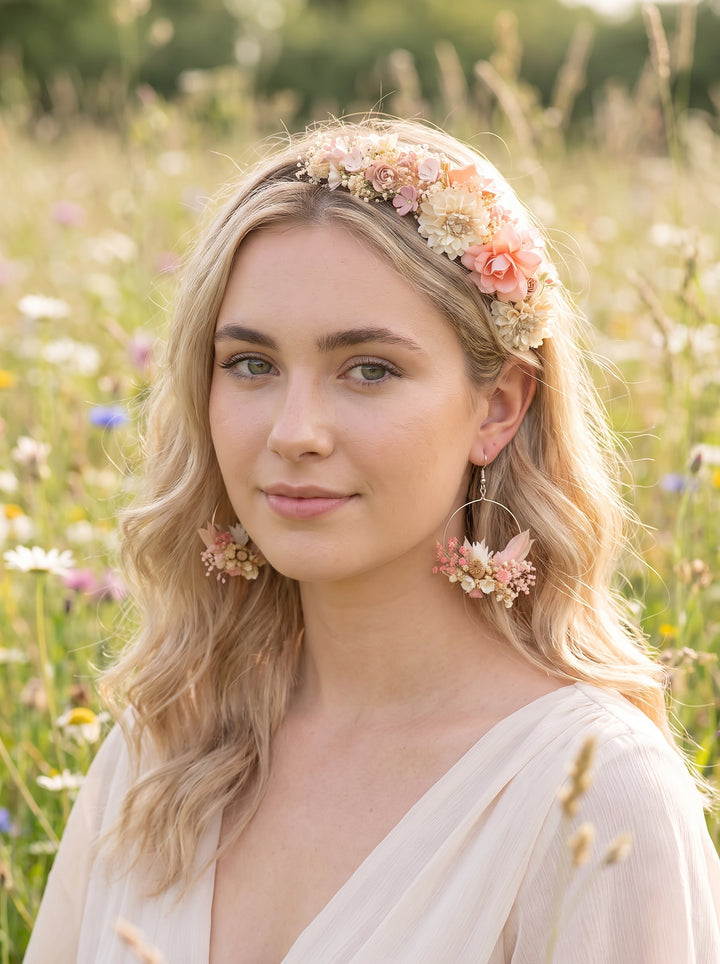 A woman with blonde wavy hair wearing a floral headband and matching dangling earrings, standing in a sunlit field of wildflowers.