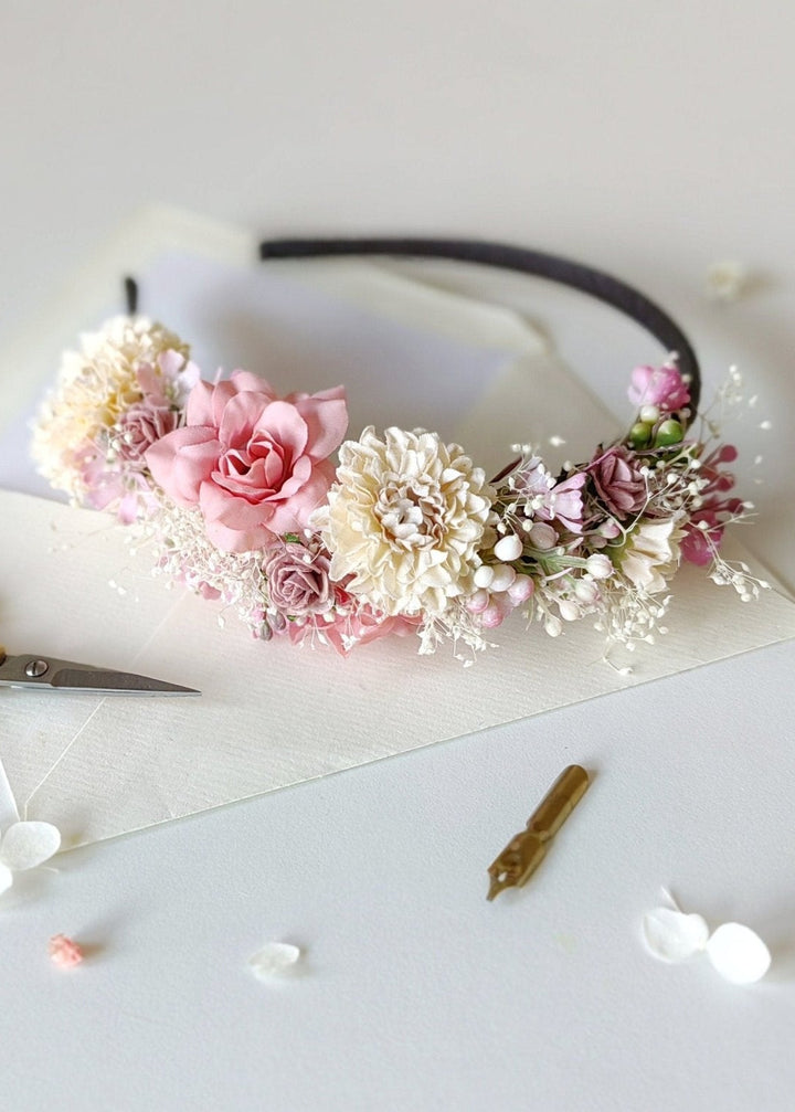A floral headband with pink and cream flowers on a black band, styled on a white surface with scissors, a small gold hair pin, and scattered flower petals.