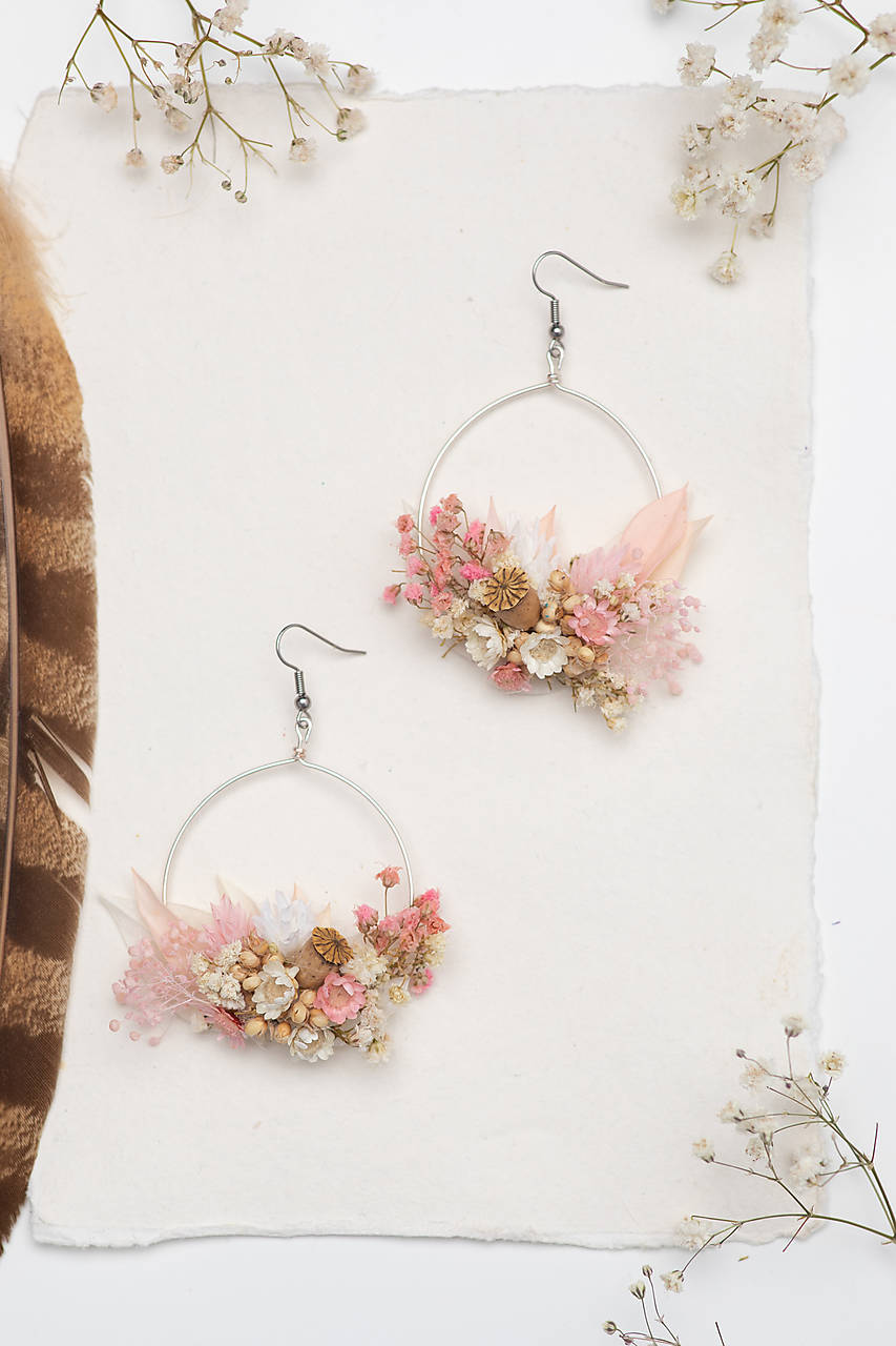 A pair of large silver hoop earrings decorated with pink and cream dried flowers, displayed on a textured white paper background with decorative baby's breath and a brown feather.
