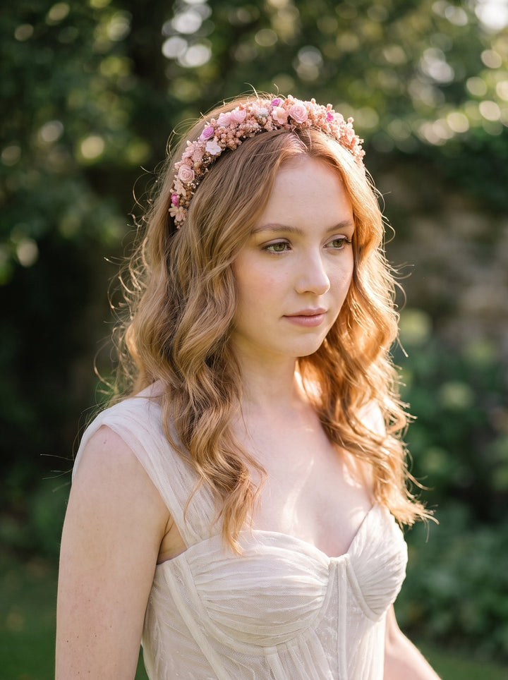 A woman outdoors wearing a delicate pink floral crown headband and a light cream sleeveless dress, with a soft blurred green garden background.