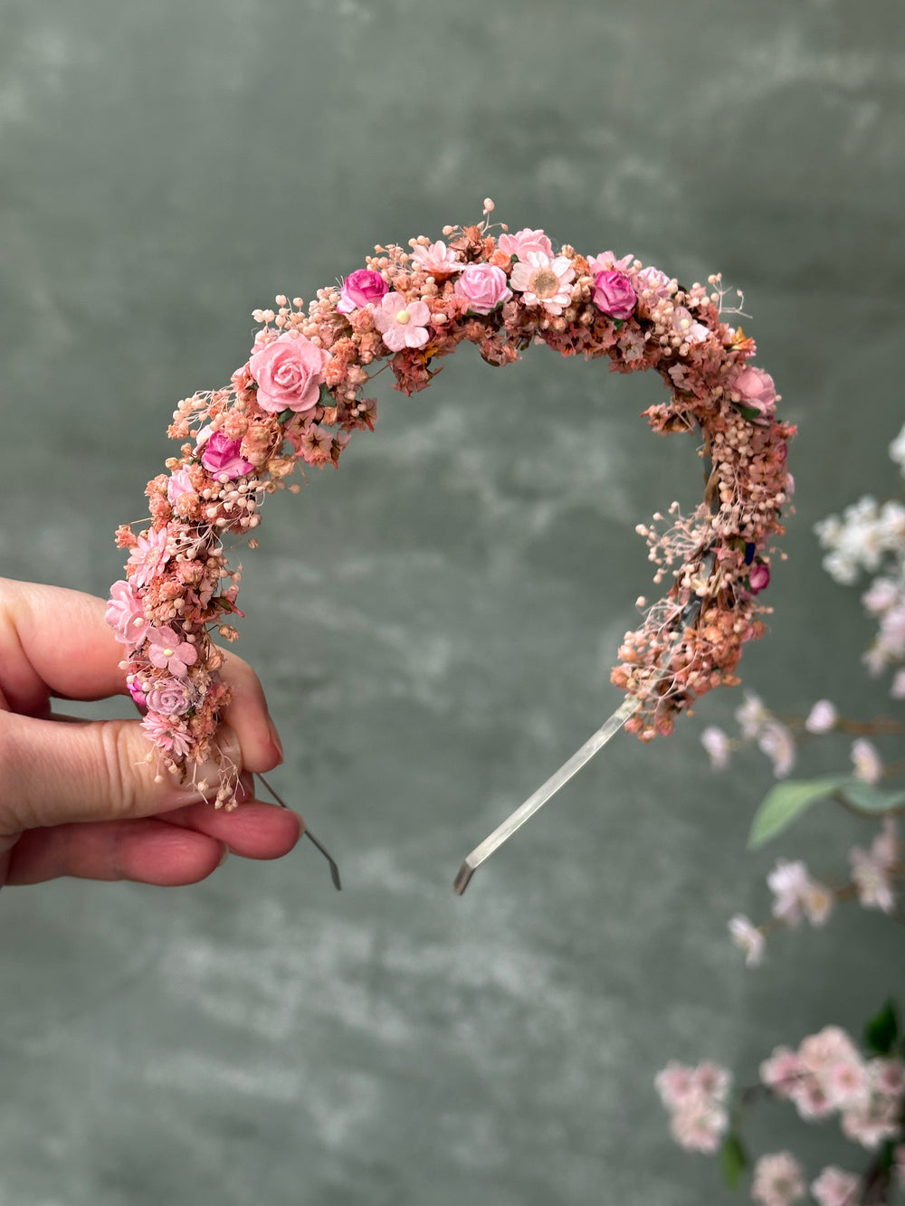 A hand holding a delicate crescent-shaped headband decorated with pink flowers, small blossoms, and brown filler against a soft gray background.