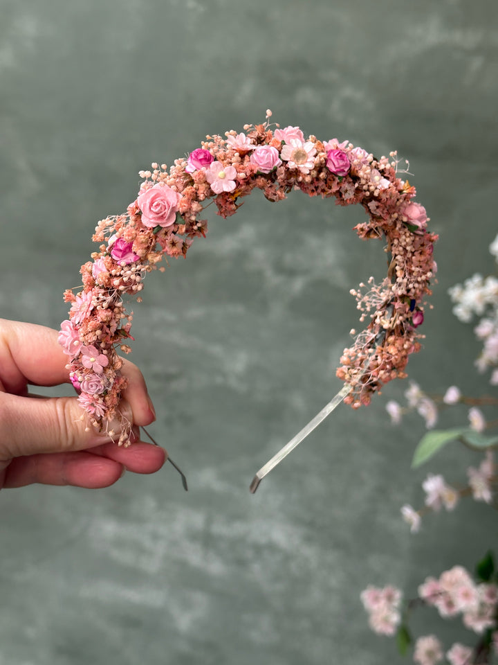 A hand holding a delicate crescent-shaped headband decorated with pink flowers, small blossoms, and brown filler against a soft gray background.