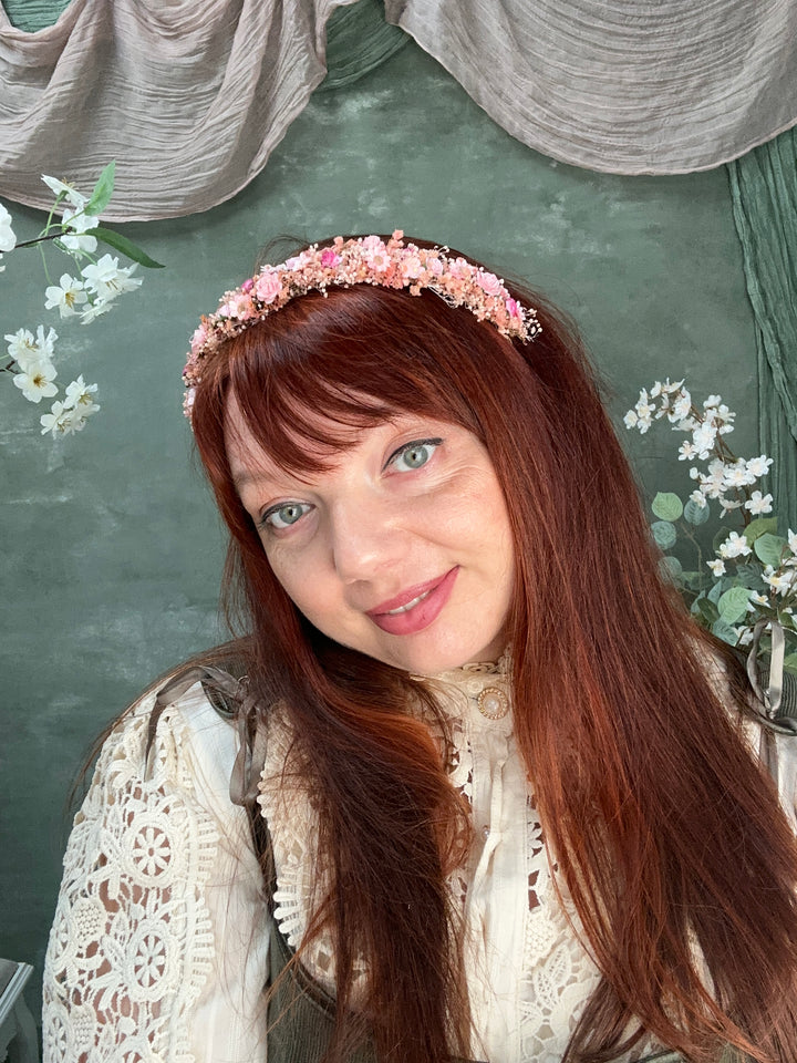 A woman with long reddish-brown hair wearing a pink floral headband and a cream lace top, photographed in a styled indoor setting with green fabric and small white flowers.