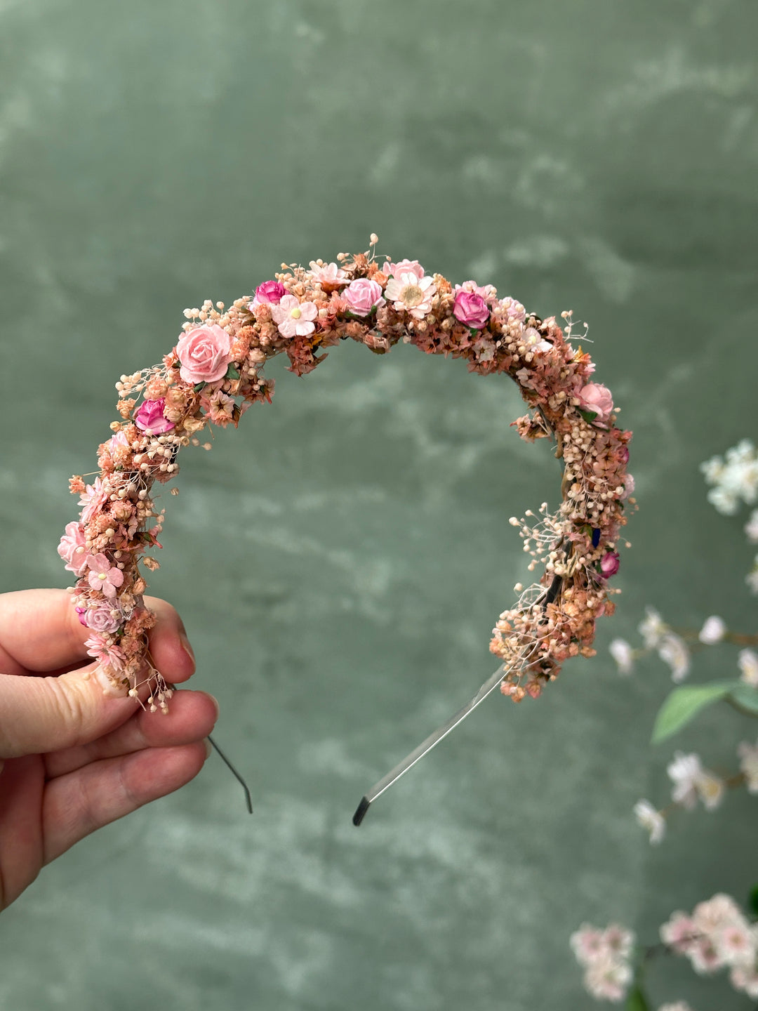 A handmade circular headband decorated with pink and peach flowers, held by a hand against a textured sage green background.