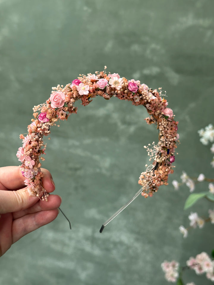 A handmade circular headband decorated with pink and peach flowers, held by a hand against a textured sage green background.