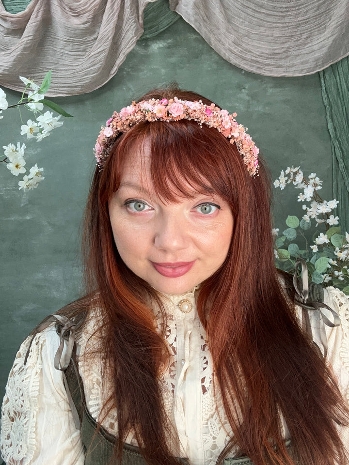 A woman with long auburn hair wearing a delicate pink floral headband and a cream lace blouse, lying on a textured dark green surface with small white flowers and gray fabric accents.