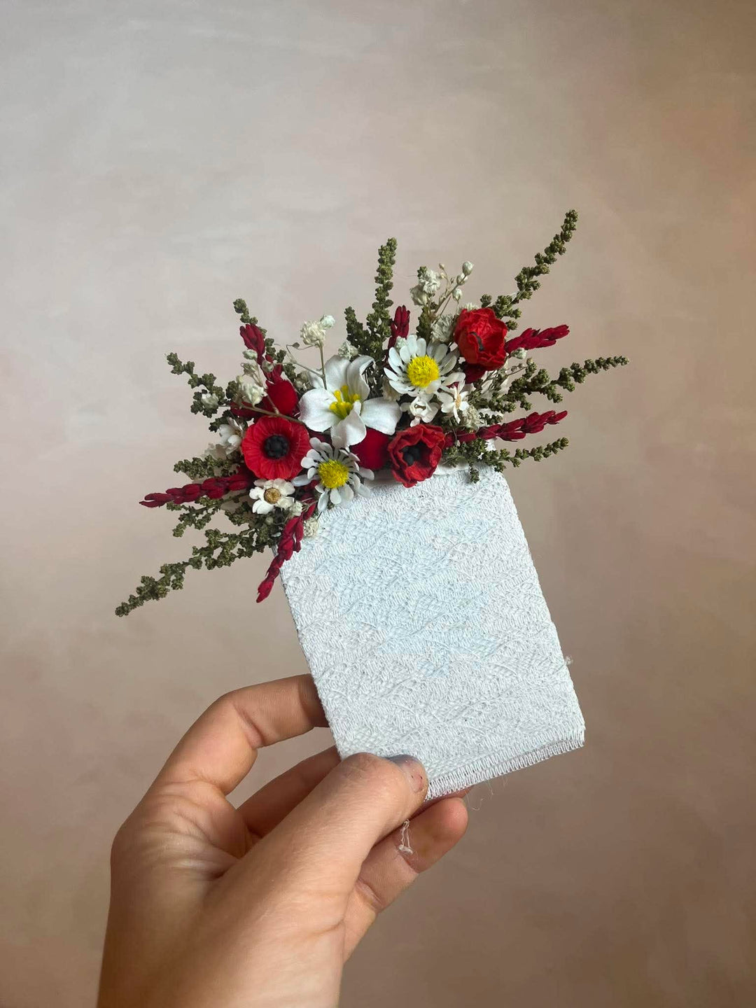 A hand holding a sboutonniere  decorated with a dried floral arrangement of red roses, white daisies, and green foliage, against a soft beige background.