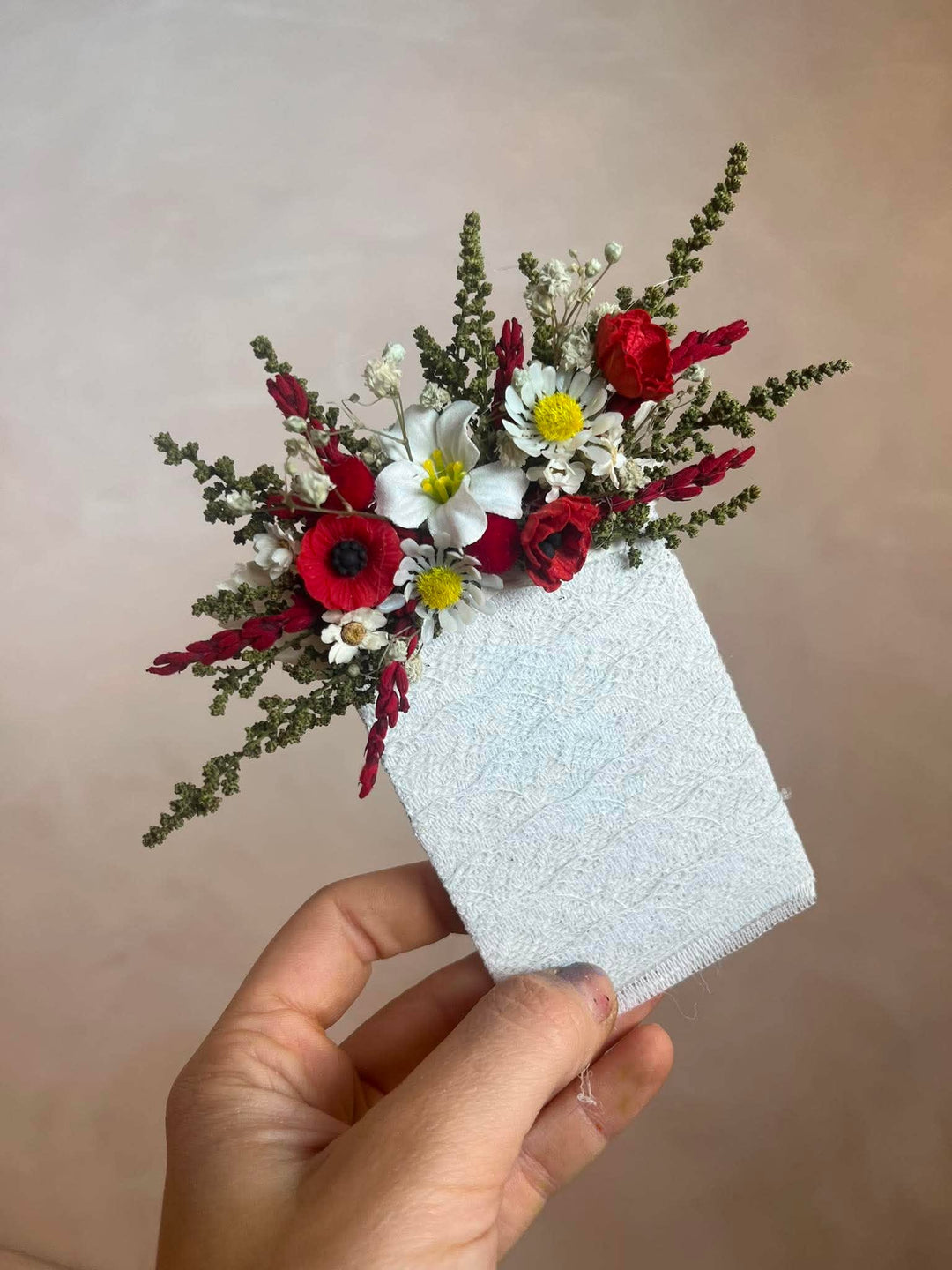 A hand holding a pocket boutonniere featuring red poppies, white daisies, and green filler stems attached to a textured white fabric base, set against a soft beige background.