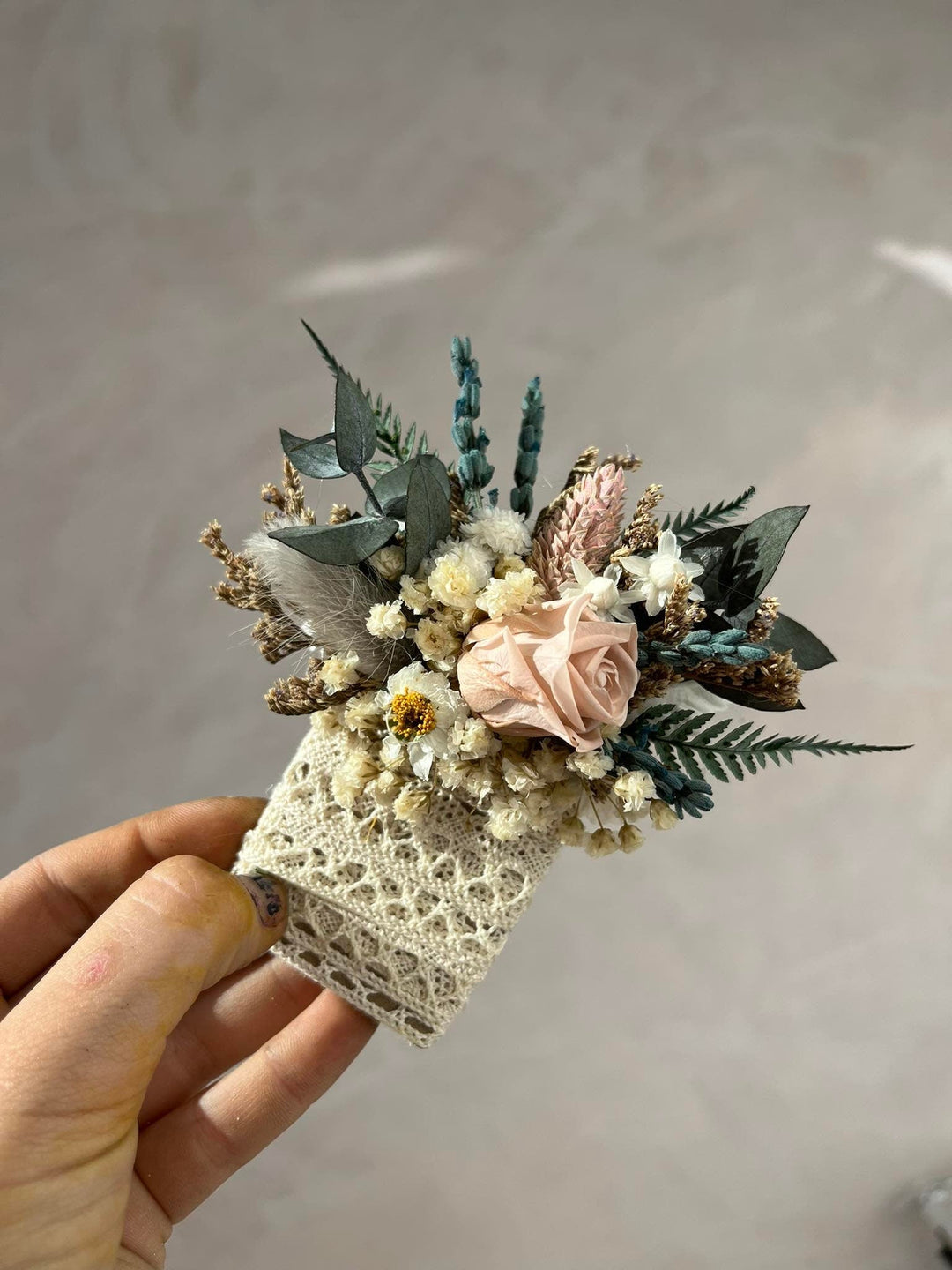 Hand holding a small floral arrangement with lace detail against a plain background