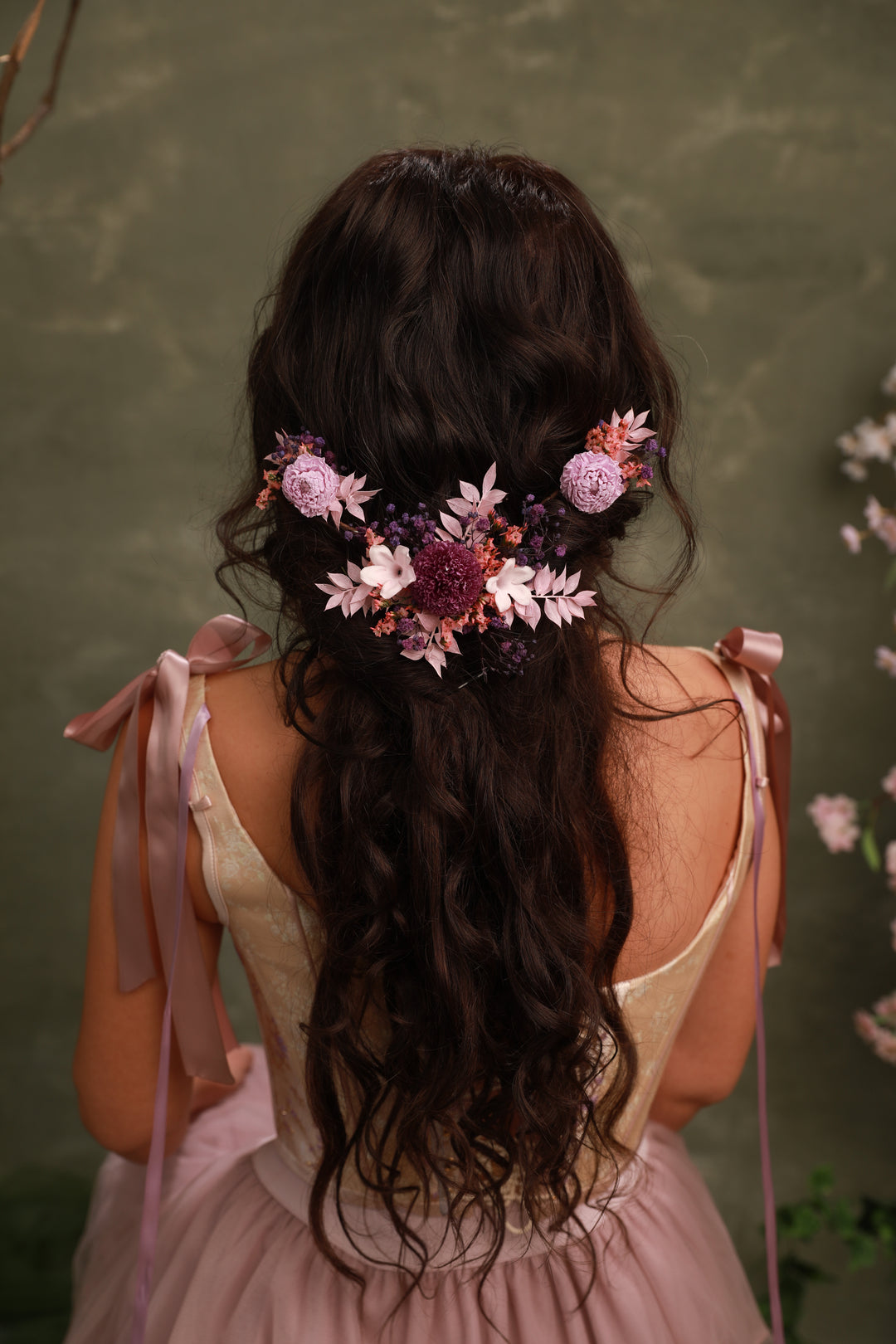 Woman with long dark hair adorned with flowers, wearing a pink dress against a green background.