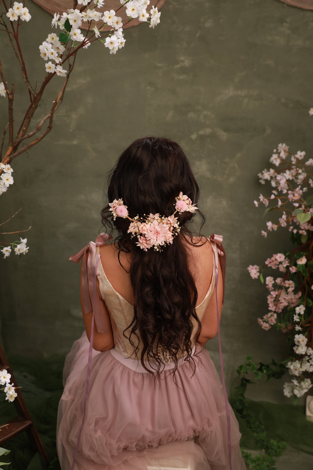 Woman with floral hair accessory sitting in a decorative setting with flowers and greenery.