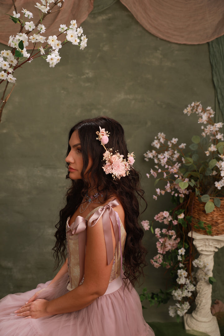 Woman in a pink dress with floral hair accessories with preserved rose and ther flowers in a decorative setting with flowers and greenery.