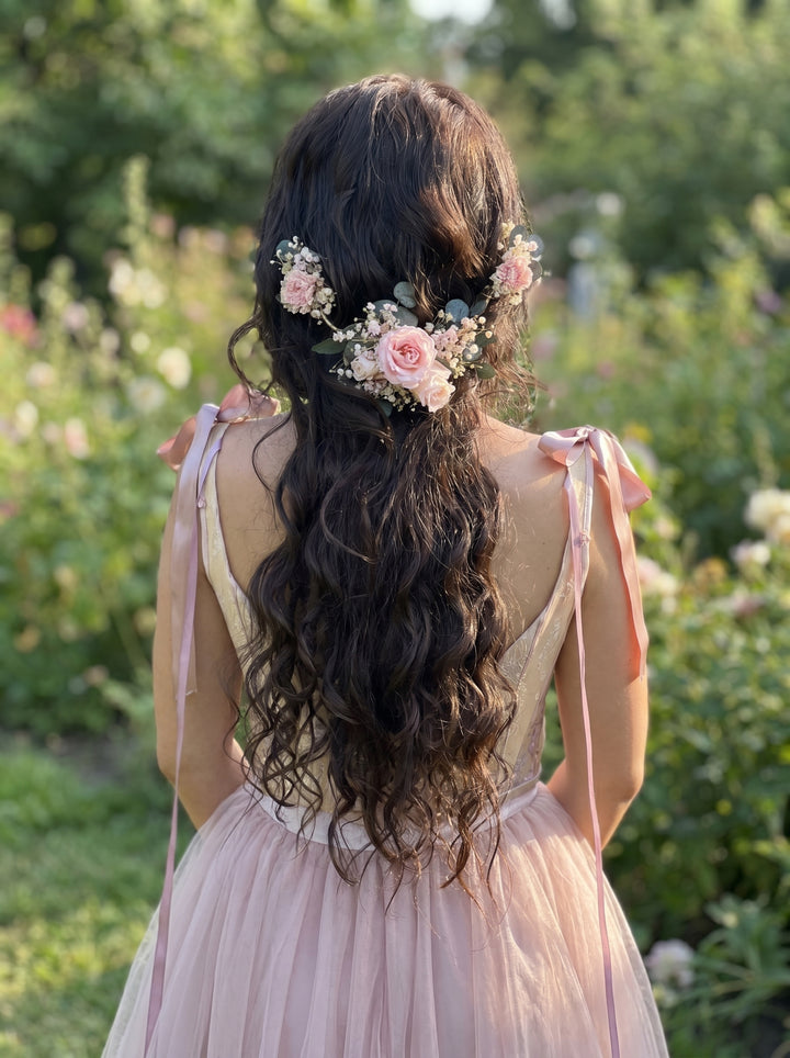 Woman with long, dark hair adorned with flower preserved hair vin in pink tones, wearing a light pink dress in a garden setting.