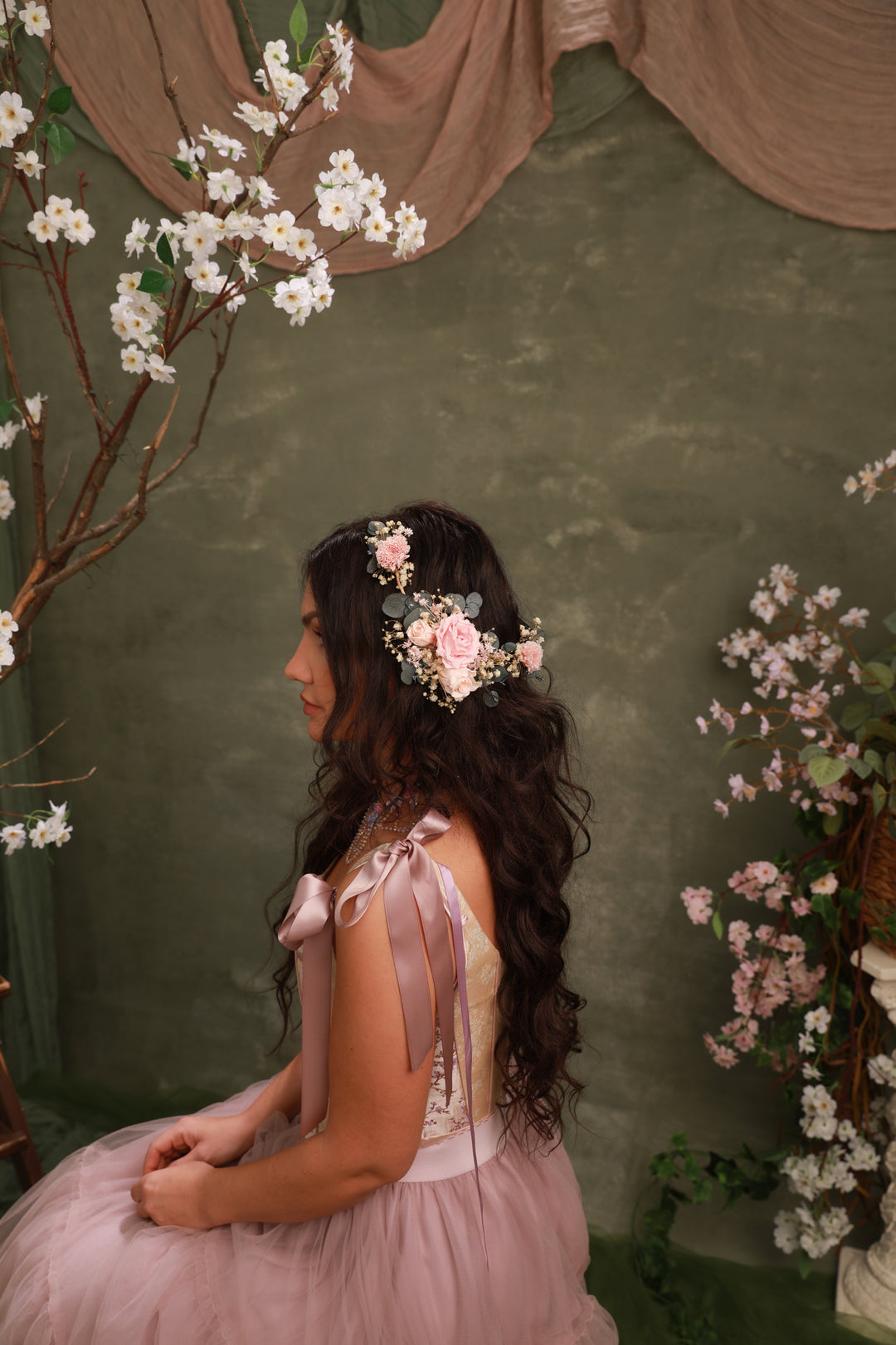Woman in a pink dress with floral headpiece sitting against a green wall with cherry blossom branches.
