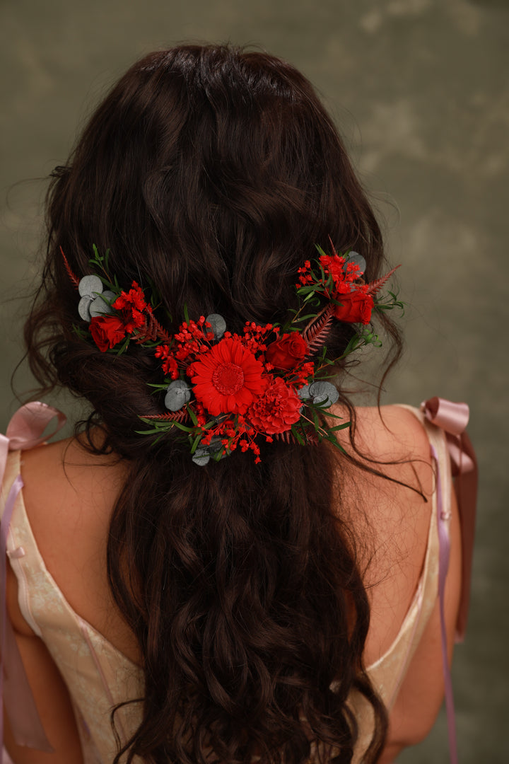 Woman with long dark hair styled in an intricate updo adorned with red flowers and greenery against a neutral background.