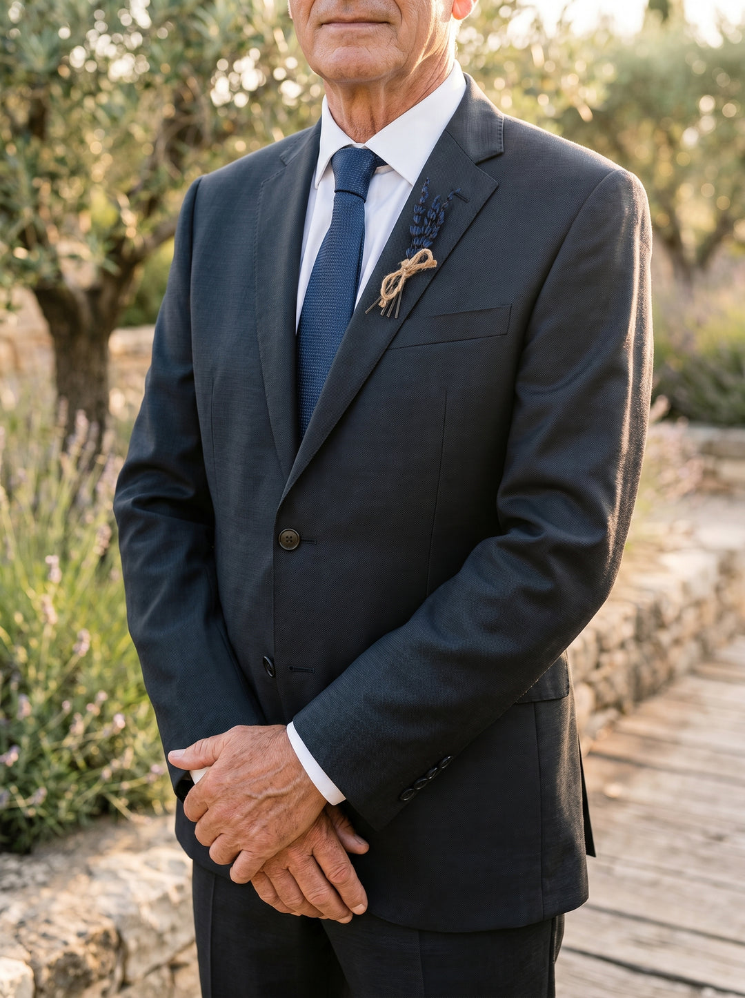 Man in a dark suit with a blue tie standing outdoors with trees and plants in the background